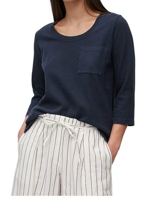 Kobieta ubrana w granatową bluzkę z rękawami do łokcia i kieszonką na piersi oraz białe spodnie z wysokim stanem, ozdobione pionowymi czarnymi paskami i materiałowym paskiem związanym w kokardę.