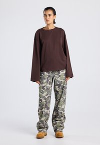 Braunes, oversized Langarmoberteil kombiniert mit locker sitzenden, camouflagemusternden grünen Hosen. Die Schuhe sind aus beigem Material und als Slipper gearbeitet.