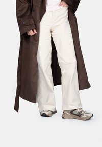 Manteau long en suède brun, pantalon large crème et baskets beiges avec des détails en maille. L'ensemble présente une coupe décontractée et des textures douces.