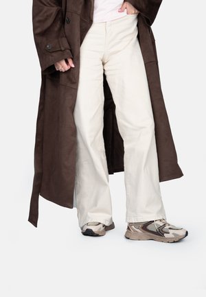 Manteau long en suède brun, pantalon large crème et baskets beiges avec des détails en maille. L'ensemble présente une coupe décontractée et des textures douces.