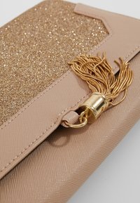 Beige käsilaukku, jossa on kimaltava kultainen kimalteinen pinta, sileä rakenne, kultaiset yksityiskohdat ja koristetupsu kullanvärisenä.