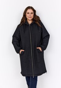 Fiatal nő hosszú, fekete steppelt kapucnis kabátban áll, elöl cipzárral és oldalsó zsebekkel, kezével a zsebében.