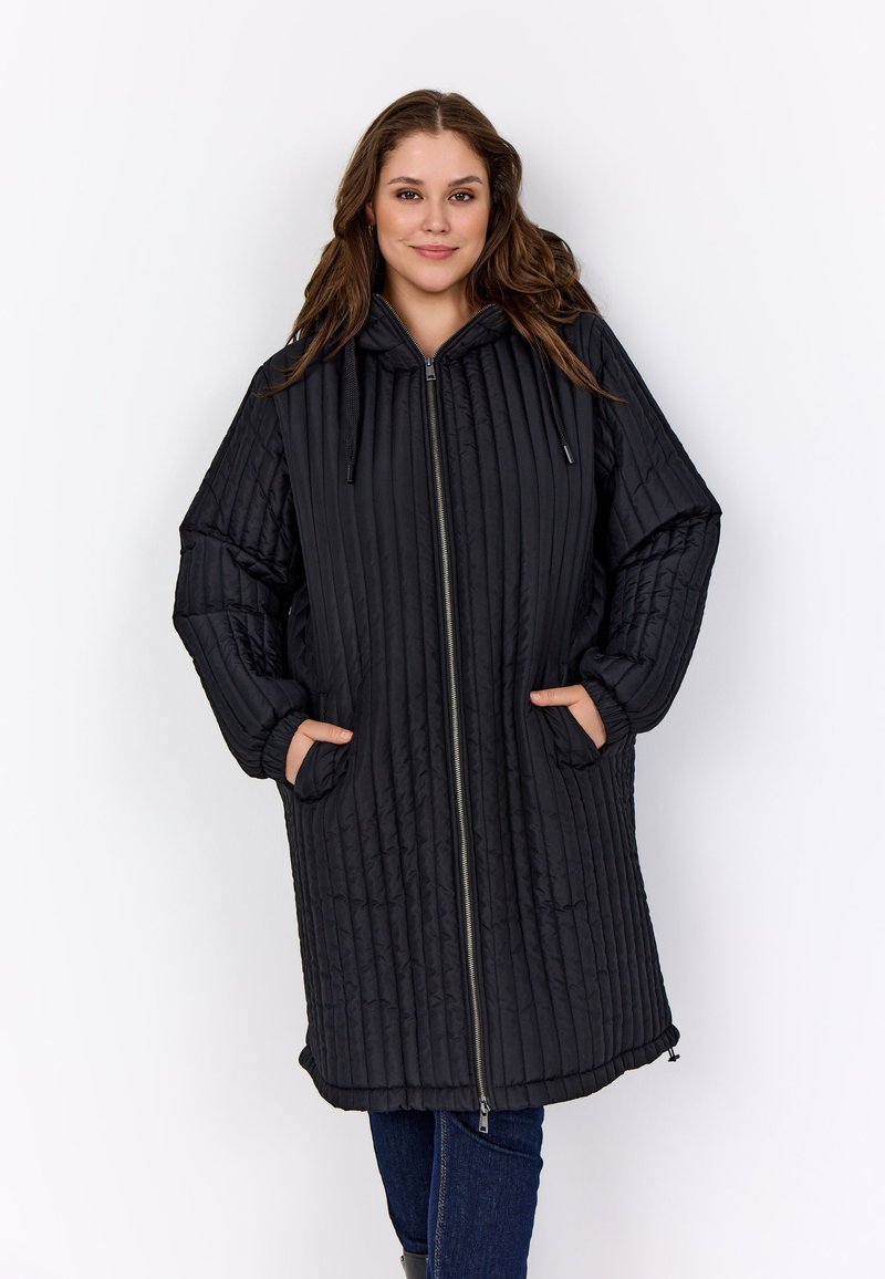 Fiatal nő hosszú, fekete steppelt kapucnis kabátban áll, elöl cipzárral és oldalsó zsebekkel, kezével a zsebében.