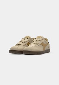 Sneakers de camurça bege com solas de borracha castanha, apresentando uma faixa lateral em tom de pele e detalhes verdes no calcanhar, atacadores à frente e gola acolchoada.