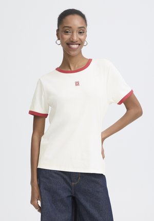 Mujer sonriente con camiseta corta de manga corta color crema con ribete rojo y pequeño logo rojo, combinada con pantalones vaqueros azul oscuro, de pie con la mano en la cadera.