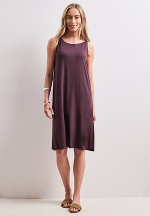 Femme debout souriante portant une robe ample sans manches violette, des bracelets argentés et des sandales plates marron sur un fond clair uni.