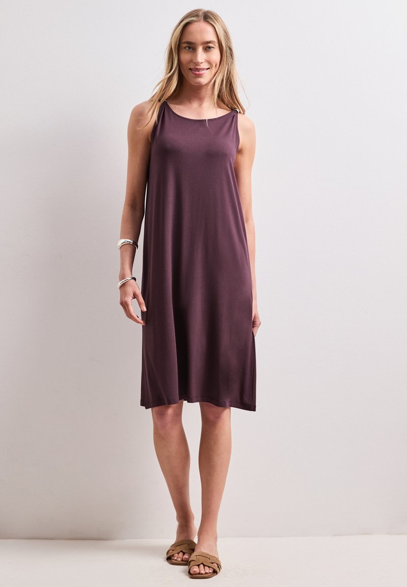 Femme debout souriante portant une robe ample sans manches violette, des bracelets argentés et des sandales plates marron sur un fond clair uni.