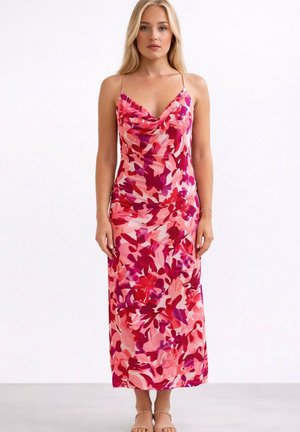 Femme portant une robe midi sans manches à fleurs roses et rouges avec de fines bretelles et des sandales à chaînes, debout devant un fond blanc.