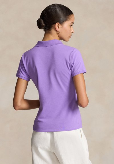 Νεαρή γυναίκα με τα μαλλιά της σε κότσο, φορά χρυσούς κρίκους στα αυτιά, ένα λιλά κοντομάνικο πόλο πουκάμισο και λευκό παντελόνι, σε προφίλ μπροστά σε μπεζ φόντο.