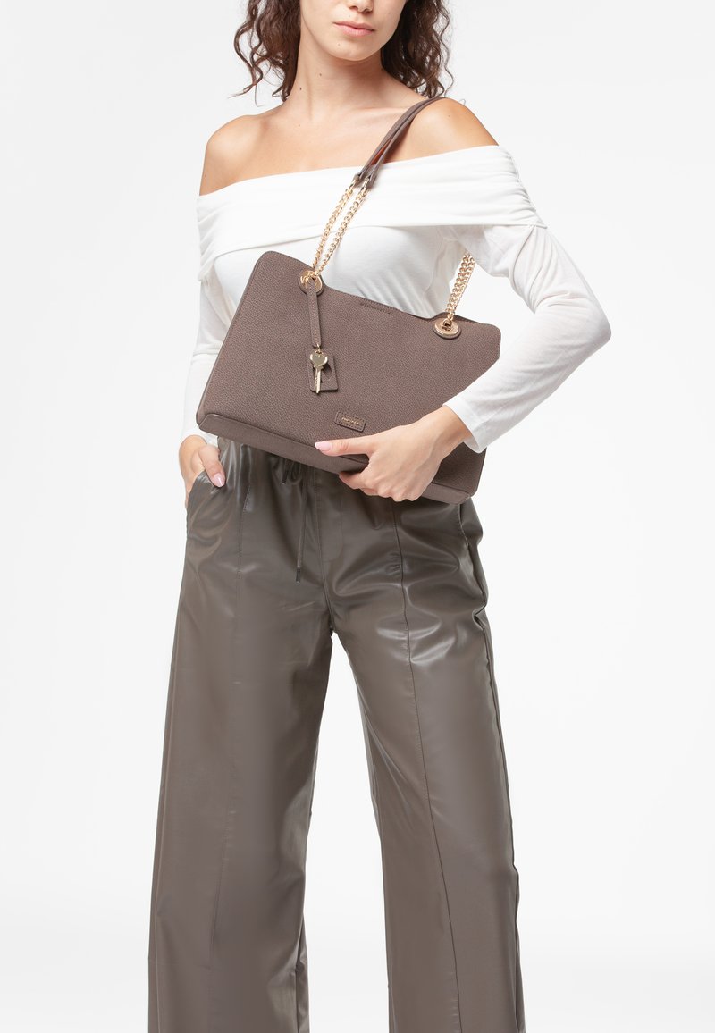 Borsa a tracolla di colore marrone con texture, dotata di catena dorata e dettaglio a chiave, tenuta da una persona che indossa un top bianco a spalle scoperte e pantaloni beige lucidi.
