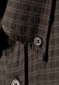 Λεπτομέρεια καφέ καρό πουκαμίσου με υφή ύφασμα, κουμπί με λουλουδένιο σχέδιο και μανίκι γυρισμένο με αντίθετη ραφή.