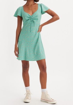 Femme portant une mini-robe vert menthe à petits motifs floraux, détail froncé à l'avant, manches volantées, chaussettes blanches et baskets montantes.