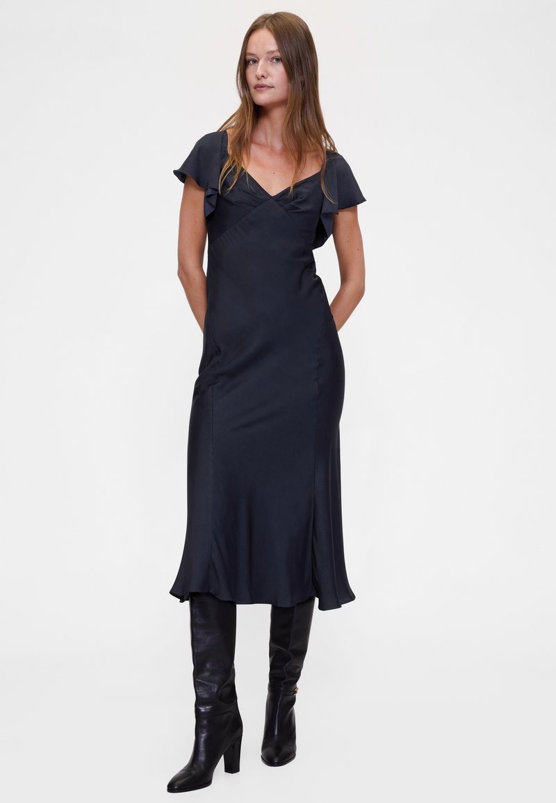 Fekete szatén ruha rövid fodros ujjakkal, V-nyakkivágással és légies sziluettel, térdig érő fekete magassarkú csizmával párosítva.