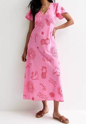 Vrouw draagt een roze midi-jurk met rode, abstracte tekeningen van ananassen, vogels, zonnen, golven en Franse woorden, gecombineerd met bruine slip-on sandalen.