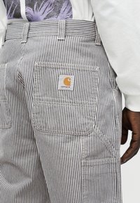 Κοντινό σε άτομο που φοράει γκρι και άσπρο ριγέ παντελόνι Carhartt με πίσω τσέπη και ορατό μανίκι λευκού πουκαμίσου μακρυμάνικου.