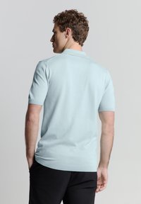 Lichtblauw poloshirt met korte mouwen van zachte stof, met een klassieke kraag en ribgebreid hem, gedragen met zwarte broek.