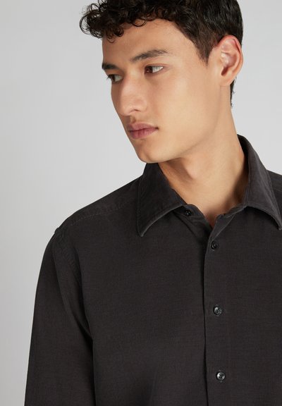 Jonge man met kort krullend haar die naar links kijkt, gekleed in een donker button-up corduroy shirt met een brede kraag tegen een neutrale achtergrond.