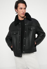 Casaco de couro preto com colarinho de pele, apresentando botões de pressão, dois bolsos frontais e um ajuste estruturado. Textura suave e design clássico.