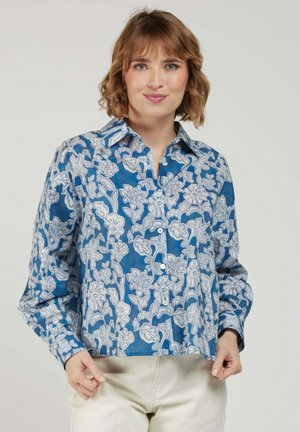 Femme portant une chemise bleue à boutons avec des motifs floraux blancs et un pantalon clair, debout devant un fond uni.