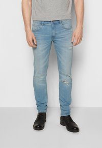 Jeans de mezclilla azul claro con un corte slim, que presentan detalles desgastados y decoloración. Combinados con botas negras. Camiseta gris visible en la parte superior.