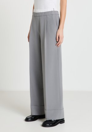 Femme portant un pantalon gris taille haute à jambes larges avec des ourlets retroussés, associé à des mocassins noirs et un haut blanc rentré.