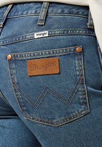 Blue denim jeans featuring a brown leather patch with "Wrangler" logo, and distinctive yellow stitching with a zigzag pattern on the back pocket.