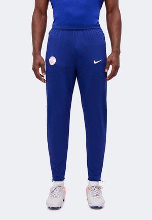 Mand iført blå Paris Saint-Germain Nike løbebukser og hvide fodboldstøvler står foran en neutral baggrund.