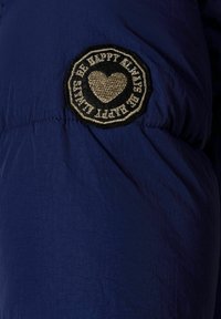 Tejido azul marino con una textura suave, que presenta un parche bordado redondo y negro con un corazón y las palabras "Siempre Sé Feliz."