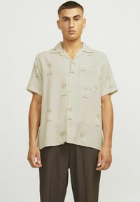 Kurzärmliges, beiges Hemd mit floralen Mustern; mit einer Brusttasche und Knöpfen, aus leichtem Stoff gefertigt. Kombiniert mit dunklen Hosen.