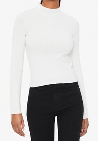 Haut blanc à manches longues côtelé avec un col montant et une silhouette ajustée, associé à un jean noir. Tissu texturé avec des lignes verticales.