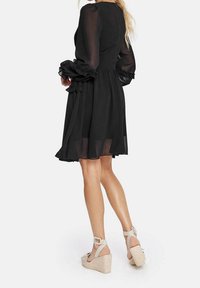 Kvinna som bär en knä-lång svart klänning med genomskinliga långa ärmar och volangdetaljer, ihop med beige kilklackar.