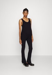 Macacão preto com nervuras, decote redondo, design justo, pernas largas e bolsos laterais. Tamancos pretos arredondados com tachas usados como calçado.