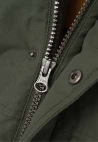 Chaqueta de tejido verde con un zipper metálico YKK, con detalles de costura y un botón a presión visible junto al zipper.
