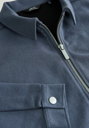 Veste zippée pour homme en bleu foncé avec une finition texturée, dotée d'un col pointu, d'une fermeture éclair argentée et d'une poche poitrine avec bouton-pression.