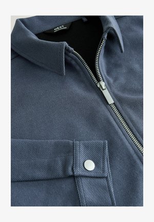 Pánska bunda na zips v tmavomodrej farbe s textúrovaným povrchom, s klasickým golierom, strieborným zipsom a náprsným vreckom so zapínaním na patentný gombík.