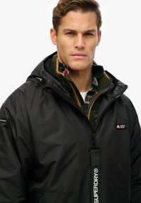 Zwarte waterdichte jas met capuchon, ritssluiting aan de voorkant en logo. Ontworpen met een binnenvoering van fleece en gelaagd over een groen geruit shirt.