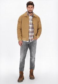 Veste en corduroy beige avec un col, fermeture à boutons, portée sur une chemise à carreaux. Un jean gris et des bottes à lacets brunes complètent la tenue.