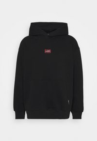 Sweat à capuche noir en coton avec manches longues, une poche avant et une petite étiquette rouge et blanche sur la poitrine. Texture douce, design décontracté.