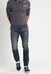 Pull à capuche gris avec cordons, jean bleu, et baskets bleu marine. Le pull présente un petit logo décoratif ainsi que des poignets et un ourlet côtelés.