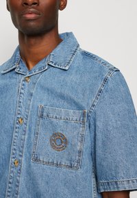 Gaiši zila džinsu krekls ar noapaļotu apkakli, īsām piedurknēm un krūšu kabatu ar apaļu izšuvumu. Pogas kontrastējošā krāsā.