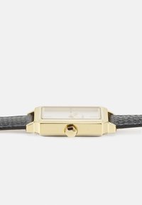 Goldfarbene rechteckige Uhr mit klarem Ziffernblatt, schwarzem strukturiertem Lederarmband und sechseckiger Krone an der Seite. Minimalistisches Design.