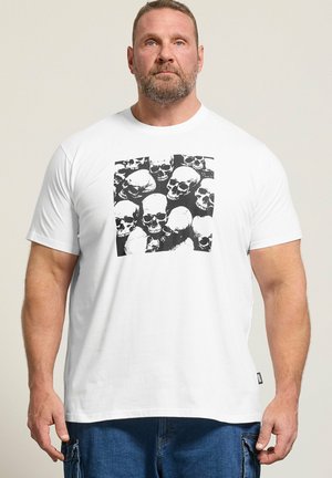 Mann mit Bart, der ein weißes T-Shirt mit schwarz-weißem Totenkopf-Cluster-Grafik trägt, kombiniert mit blauen Cargohosen, vor neutralem Hintergrund stehend.