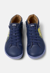 Baskets pour tout-petits de couleur bleu marine avec un graphisme de dinosaure jaune, à lacets à l'avant, fermeture éclair sur le côté et semelle de couleur claire, vues de dessus sur fond blanc.
