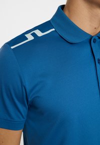 Blå pikétröja med krage, som har ett subtilt ljusblått mönster på axeln. Tillverkad av ett mjukt, lätt material.