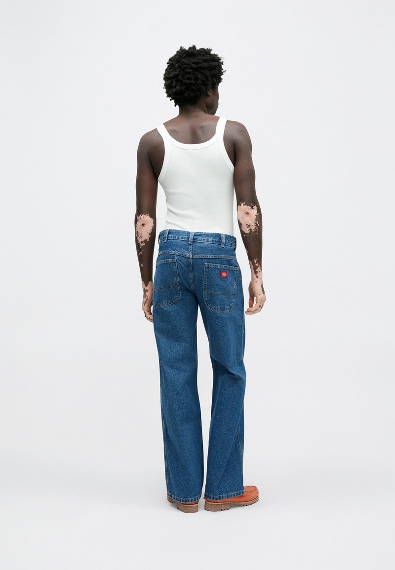 Blue denim jeans with a loose fit, featuring a classic five-pocket design and a red logo patch, paired with a white tank top.