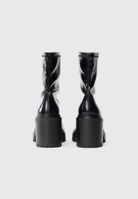 Une paire de bottes noires brillantes à mi-mollet avec des talons hauts épais, vues de l'arrière sur un fond blanc uni.