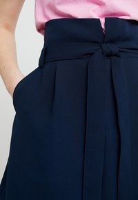 Mörkblå hög midja byxor med ett brett bälte, utrustad med två sidofickor och diskreta veck för extra textur och designdetaljer.