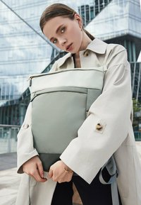 Un sac à dos gris clair au fini lisse, avec une poche avant à fermeture éclair et des bretelles réglables, tenu par une personne portant un trench-coat beige.