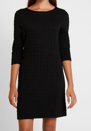 Femme portant une robe noire texturée longueur genou avec des manches trois-quarts et un col rond, debout devant un fond clair uni.