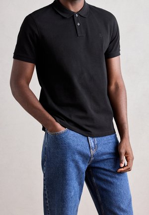 Mann trägt ein schwarzes, kurzärmliges Poloshirt und blaue Jeans, mit einer Hand in der Tasche und der anderen locker an der Seite.